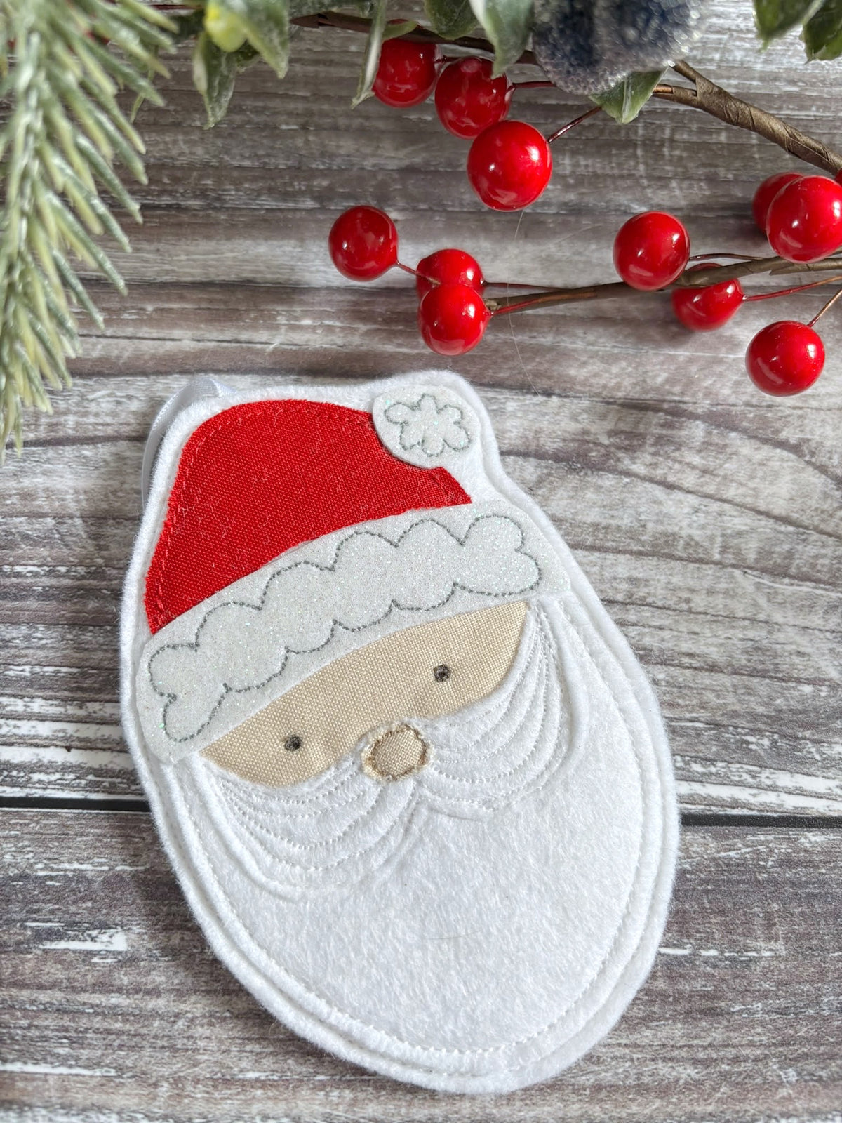 A handmade felt Father Christmas face decoration styled on a rustic gray wooden surface with red berries and green pine branches.